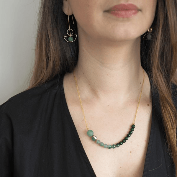 A necklace made up of labradorite, aventurine and amazonite beads in varying shade of green on a brass chain, shown on a white model who wears a black v-neck top and brass and aventurine earrings.