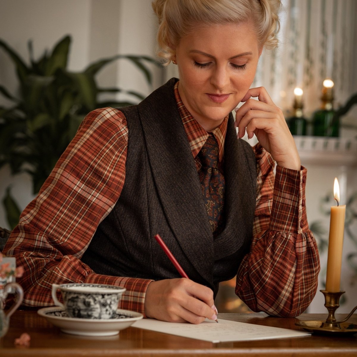 Designer Emmy from Emmy Design Sweden wear the Bishopess Blouse in rusty plaid, a long sleeved vintage inspired blouse, underneath a waistcoat with a dark printed tie. She sits at a vintage desk writing
