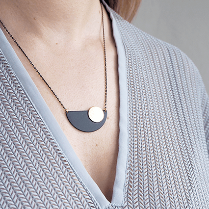 An Art Deco inspired necklace made up of a black crescent with a brass circle set off centre, shown against white skin and a v-neck top