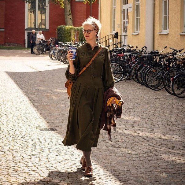 Emmy Design Sweden designer Emmy wears the Green Gable Dress in forest green whilst strolling down a cobbled street carrying a coffee