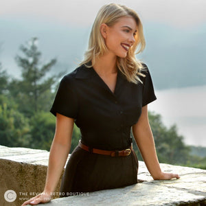 A white model with blonde shoulder length hair wears a black blouse tucked into brown high waisted trousers with a tan leather belt