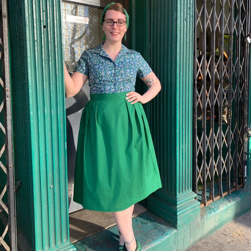 Vintage style girl wears Liberty print floral blouse in blue and green tucked into a green a-line skirt with pleats at the sides.