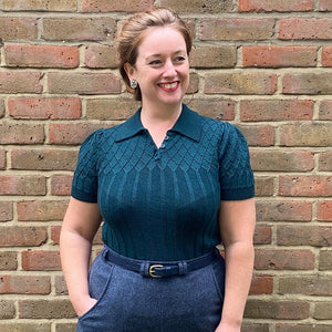 Mary, a white woman with strawberry blonde hair pulled back off her face, stands straight on to the camera wearing a bottle green vintage style knitted jumper with collar tucked into blue herringbone high waist trousers worn with a navy leather belt