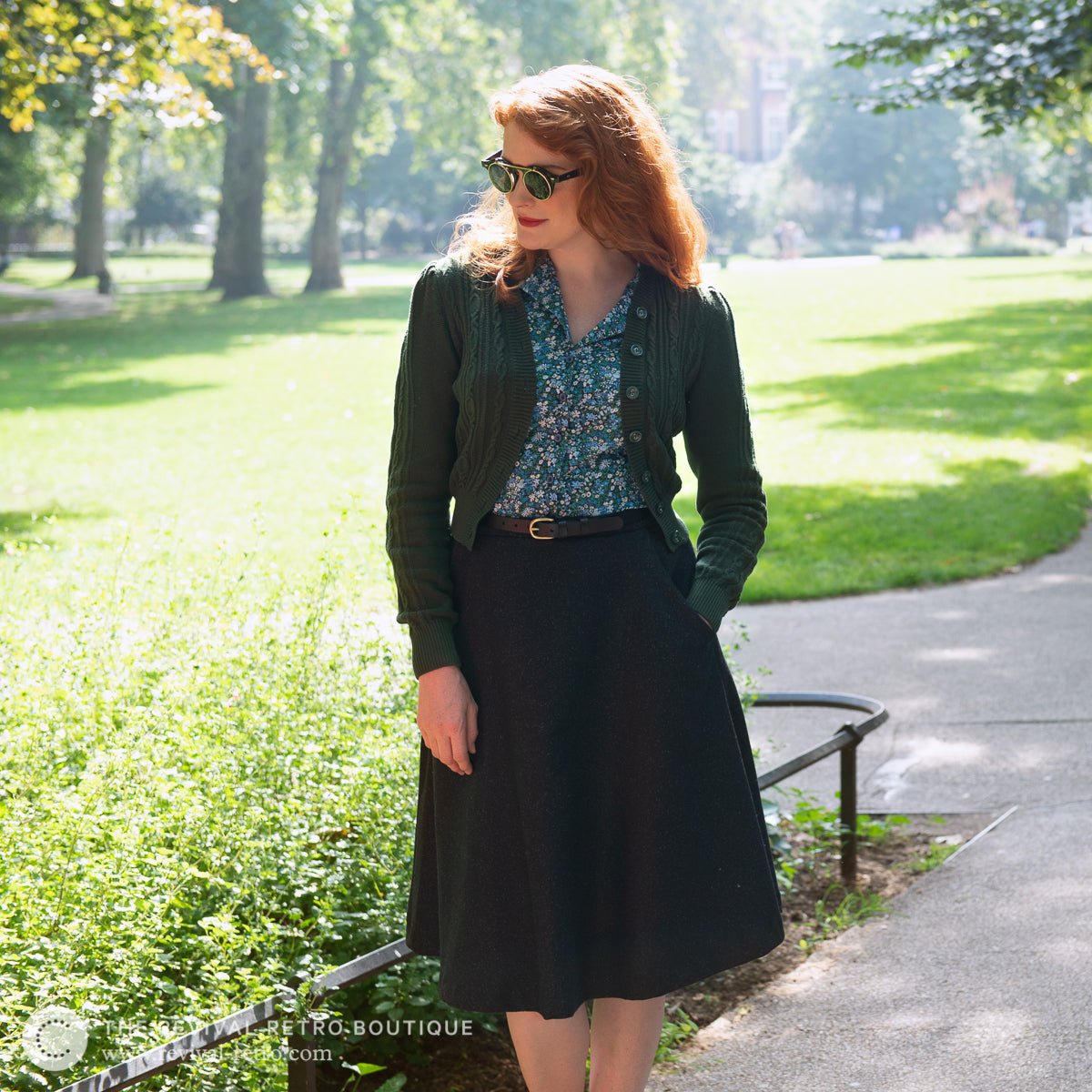 1940s inspired vintage outfit made up of a cropped knitted Emmy Cardigan in pine green, Liberty print blouse and charcoal a-line skirt, all worn by a white model with ginger hair and 1940s round sunglasses