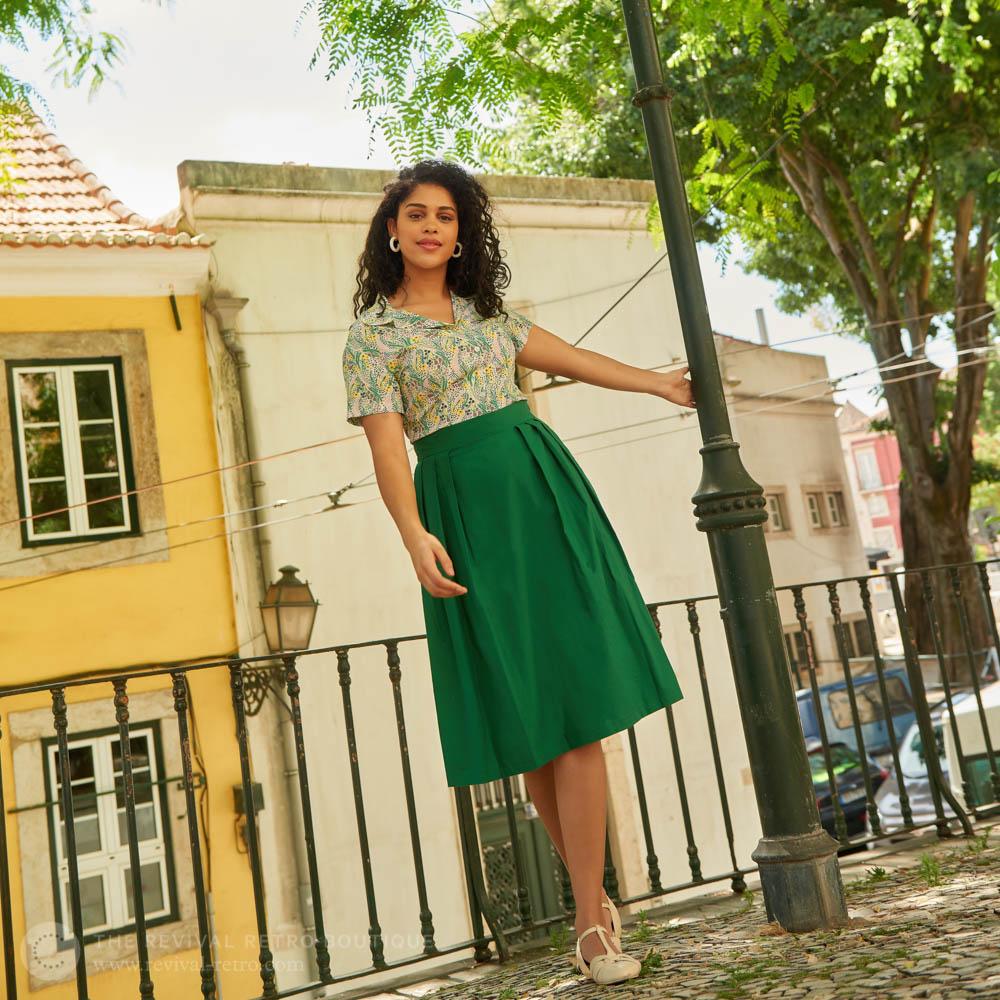 Model wears Liberty print blouse tucked into a green a-line skirt pleats
