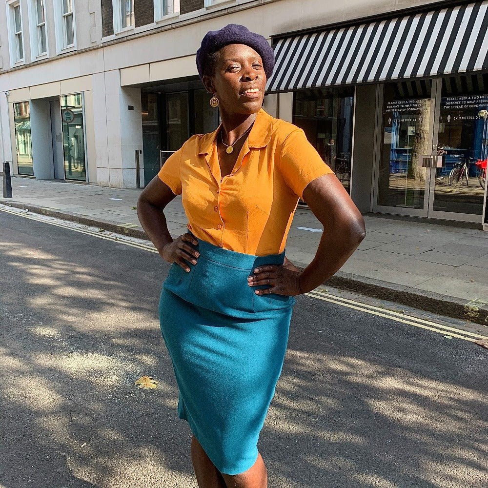Black model Anna wears a vintage style outfit comprising of navy beret, mustard shirt with round collar tucked into a high waisted teal pencil skirt. She stands with her hands and her hips.