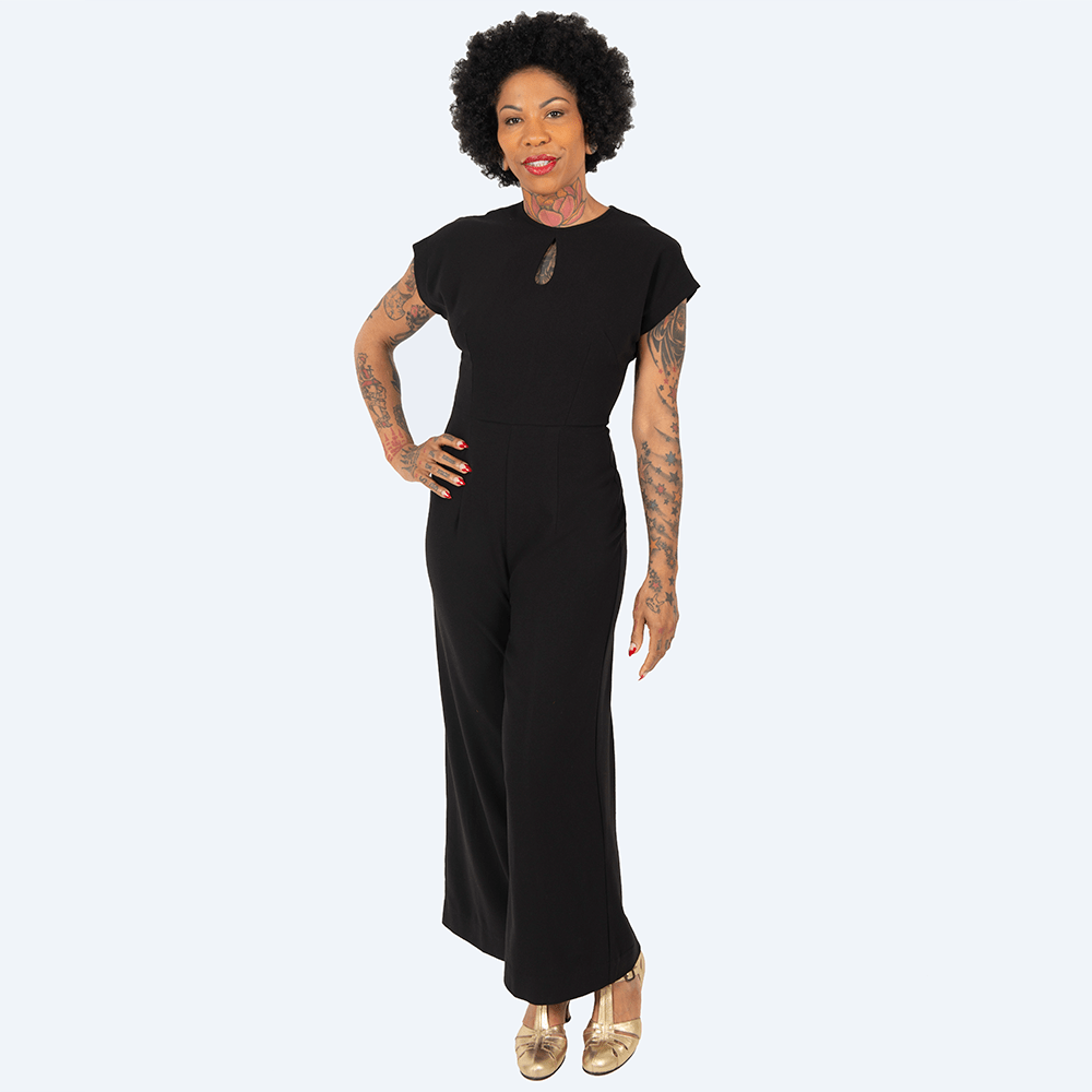 Full length view of Retro Style Trouser Suit, Mayfair Jumpsuit in black, as worn by a model who is a woman of colour with natural hair who stands with her right arm on her hip looking straight at the camera.