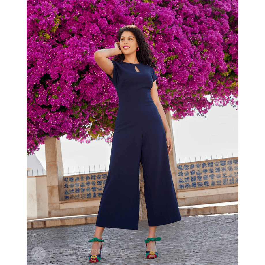 Full length picture of Retro Style Trouser Suit, Mayfair Jumpsuit in navy as worn by a professional model who is a woman of colour who is tucking her long curly hair behind her ear whilst looking off to her right and smiling.