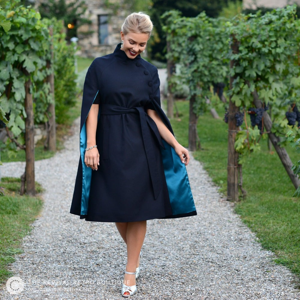 Blonde lady walks towards camera wearing navy knee length cape, vintage inspired jewellery and shoes whilst showing the kingfisher blue lining of the cape