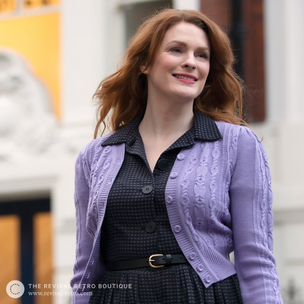 A white mid sized model with ginger hair wears a dark check shirt dress with a lilac cardigan open over the top and a black leather belt around her waist
